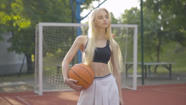 Potret menarik bijaksana wanita muda berdiri dengan bola di bawah sinar matahari. Pendekatan kamera untuk pemain basket Kaukasia yang indah berdiri di taman bermain dan tersenyum. — Stok Video