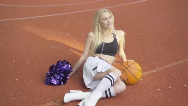 Portrait de joyeux jeune pom-pom girl mince assis sur le court extérieur avec basket-ball et pom pom pom. Souriant blond Femme caucasienne reposant sur le terrain de sport de basket-ball au soleil le jour de l'été. — Video