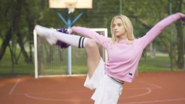 Kendine güvenen ponpon kız dışarıda bacağını kaldırıyor ve ponponları sallıyor. Basketbol sahasında antrenman yapan güzel sarışın bir kadının portresi. Çekici Kafkas spor dansçısının antrenmanı. .