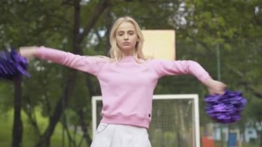 Açık sahada ponpon kızlarla dans eden kendine güvenen sarışın bir kadın. Pozitif özgüvenli beyaz amigo kız portresi güneşli bir yaz gününde dışarıda dans ediyor ve kameraya gülümsüyor..