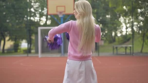 Fröhliche Cheerleader, die sich kameraschwingenden Pompons zuwenden. Porträt einer schönen jungen kaukasischen Frau, die auf einem Spielplatz im Freien posiert und lächelt. Glückliche blonde Sportlerin auf Sportplatz. — Stockvideo