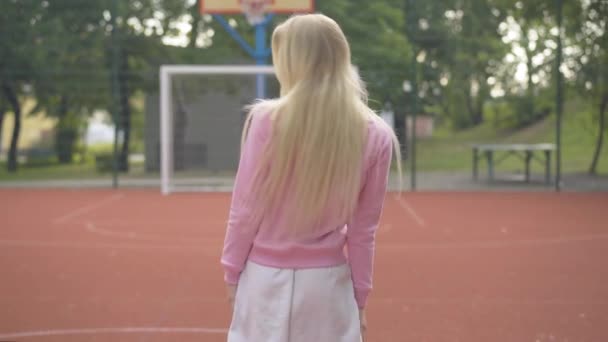 Donna bionda magra che si gira verso la telecamera e sorride. Ritratto di giovane sportiva caucasica felice in posa sul campo da basket all'aperto. fotocamera si avvicina a allegro bella femmina giocatore. — Video Stock