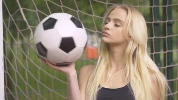 Primer plano de la joven portera posando con pelota en el patio al aire libre. Retrato de la hermosa deportista caucásica confiada sonriendo a la cámara en la red de fútbol. — Vídeos de Stock