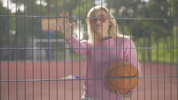 Portret pięknej sportsmenki pozującej z piłką do kosza na siatce ogrodzenia w promieniach słonecznych. młody pewny biały kobieta z blond włosy stojący w sunlight na letni plac zabaw. — Wideo stockowe