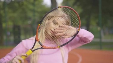 Güneşli yaz gününde tenis raketiyle poz veren neşeli sarışın kadının yakın çekimi. Dış sahada durup kameraya gülümseyen güzel beyaz bir oyuncunun portresi..