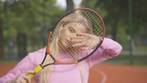 Közelkép egy vidám szőke nőről, aki teniszütővel pózol a napsütéses nyári napon. Portré gyönyörű fiatal kaukázusi játékos áll a szabadtéri pályán, és mosolyog a kamera. — Stock videók