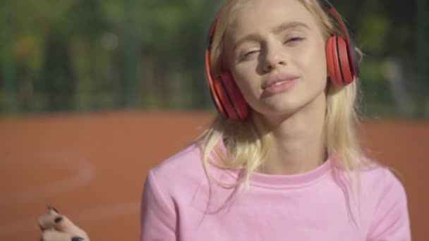 Primer plano de la joven mujer caucásica feliz en auriculares cantando y sonriendo en el soleado día de verano al aire libre. Retrato de hermosa dama rubia divirtiéndose en el campo de deportes. — Vídeo de stock