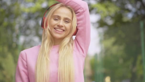 Retrato de la joven rubia feliz bailando en el soleado parque de verano al aire libre. Alegre dama caucásica disfrutando de la música en los auriculares. Alegre encantadora mujer descansando en la luz del sol. — Vídeo de stock