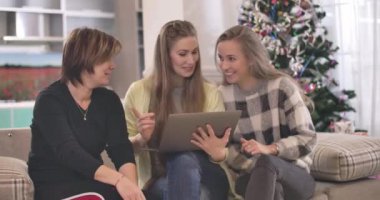 Kanepede oturan ve dizüstü bilgisayar kullanan üç beyaz kadın. Gülümseyen arkadaşların Noel 'i evde geçirdikleri portresi. Yılbaşı arifesi, tatil sezonu. Sinema 4k ProRes Merkezi.