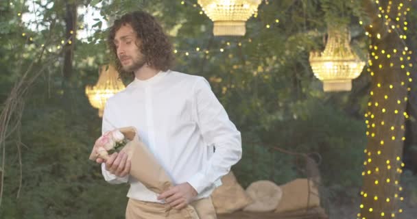 Το μεσαίο πορτραίτο του νευρικού Καυκάσου άνδρα που στέκεται με μπουκέτο τριαντάφυλλα και ελέγχει το χρόνο. Λατρεύω τον ρομαντικό σγουρό γκόμενο που περιμένει την κοπέλα του το βράδυ του καλοκαιριού. Κινηματογράφος 4k ProRes HQ. — Αρχείο Βίντεο