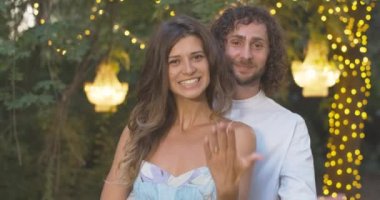 Yüzüklerini gösteren ve kameraya gülümseyen mutlu beyaz bir çiftin portresi. Olumlu nişanlın ve gelinin yaz akşamı nişanı var. Aşk ve aşk kavramı. Sinema 4k ProRes Merkezi.