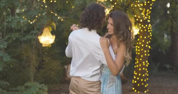 Middle shot of happy young couple dancing outdoors on summer evening. Cheerful Caucasian man and woman dating with romantic lights at the background. Slow dance of loving partners. Cinema 4k ProRes HQ — Stock Video