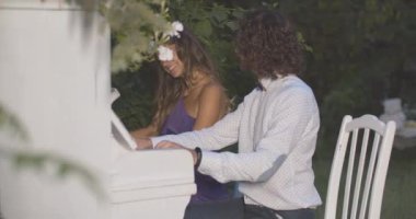 Akşam bahçesinde piyano çalan tutkulu çiftin yan görüntüsü. Şık beyaz erkek ve kadın müzisyenlerin alacakaranlıkta performans sergiledikleri portre. Sinema 4k ProRes Merkezi.