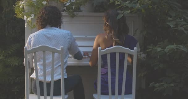 Alacakaranlıkta yaz bahçesinde piyano çalan mutlu beyaz çiftin arka görüntüsü. Genç bir adam ve kadın açık havada birlikte gösteri yapıyorlar. Sinema 4k ProRes Merkezi. — Stok video