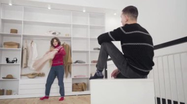 Erkek arkadaşına kıyafet gösteren mutlu ve genç bir kadın. Adam onaylıyor. Neşeli beyaz çiftin geniş açılı portresi Noel kutlaması için hazırlanıyor..