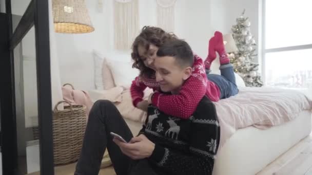 Mooie vrolijke vrouw knuffelen jonge man met behulp van smartphone. Portret van een jong blank echtpaar dat geniet van kerstvakantie met nieuwjaarsboom op de achtergrond. Liefde en romantiek. — Stockvideo