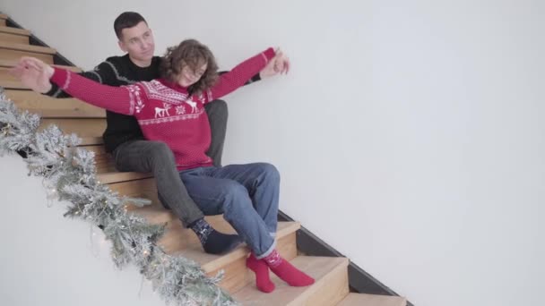 Älskade man och kvinna sitter på trappor med utsträckta händer och kramar. Porträtt av glada romantiska kaukasiska par njuter av julen hemma. Nytt år för leende pojkvän och flickvän. — Stockvideo