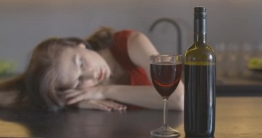 Önünde kırmızı şarap olan bir şişe ve cam. Arka planda depresif bir kadın yatıyor. Sarhoş, Kafkasyalı kadın, yalnız bir akşamı alkolle geçiriyor. Sinema 4k ProRes Merkezi.