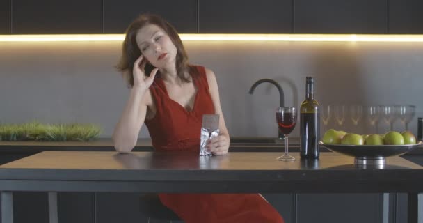 Prachtige depressieve volwassen vrouw in rode jurk zittend in de keuken en chocolade etend. Portret van een mooie blanke dame die een eenzame avond thuis doorbrengt. Depressie concept. Cinema 4k ProRes Hoofdkwartier. — Stockvideo