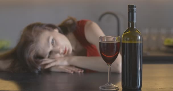 Garrafa e copo com vinho tinto na frente com mulher deprimida borrada deitada na mesa ao fundo. Bêbado infeliz caucasiano senhora passar a noite solitária com álcool. Cinema 4k ProRes HQ. — Vídeo de Stock