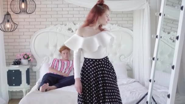 红头发的年轻白人男人躺在床上，看着美丽的女人在镜子里欣赏她的倒影。迷人的夫妇准备出去玩。关系概念. — 图库视频影像