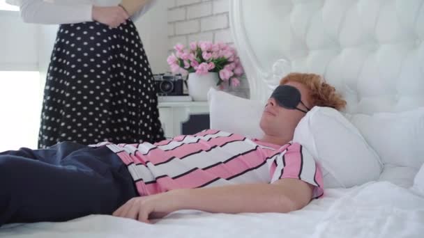 Shocked young redhead man waking up as unrecognizable woman standing next to bed with rolling pin. Portrait of guilty boyfriend and angry girlfriend in bedroom. Conflict concept. — Stock Video