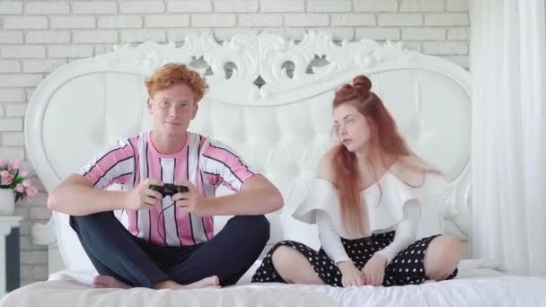 Wanita yang kesal melihat pria berambut merah bermain video game. Potret pasangan muda Kaukasia duduk di tempat tidur. Pacar tampan yang kecanduan mengabaikan pacar yang cantik. — Stok Video