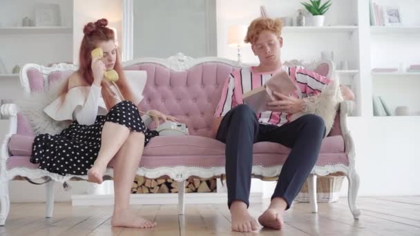 红头发女人在复古电话上用男人读的书的方式拨打号码。一对快乐的白人年轻夫妇坐在客厅沙发上的画像。休闲和生活方式。复古概念. — 图库视频影像
