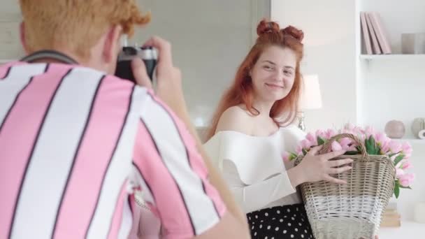 Portret uśmiechniętej rudej kobiety trzymającej kosz z tulipanami jako mężczyzna robiącej zdjęcia przed kamerą. Czarująca, biała dziewczyna udająca chłopaka, który fotografuje prezent. Pojęcie miłości. — Wideo stockowe