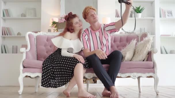 Szerokie ujęcie szczęśliwej młodej pary siedzącej na kanapie i robiącej selfie aparatem retro. Portret białej rudej kobiety i mężczyzny cieszących się weekendami w domu. Koncepcja vintage. — Wideo stockowe