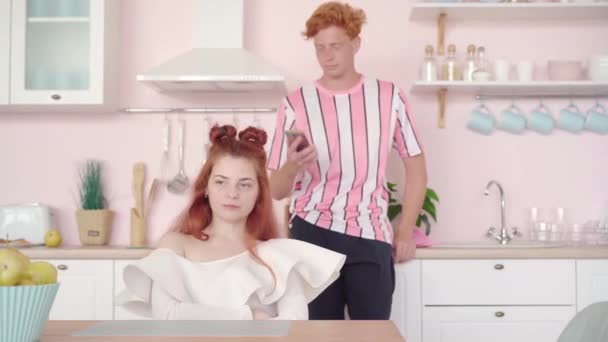 남자가 뒤에서 전화로 통화하는 동안빨간 머리 여자가 분홍색 부엌에 앉아 있는 것이 행복하지 않았다. 백인 여자 친구의 초상화는 남자 친구에게 무시당하고 있어요. 의사소통 문제. — 비디오