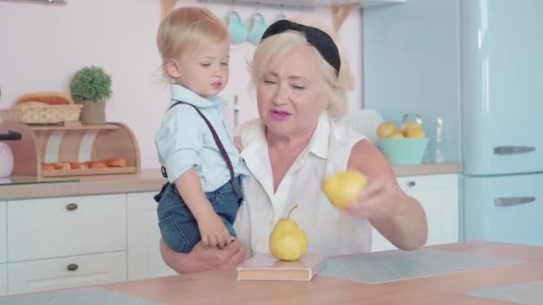 Portrait of beautiful Caucasian grandmother offering pears to cute little grandson. Happy senior woman enjoying weekends with pretty boy at home in kitchen. Aging and happiness concept. — Stock Video