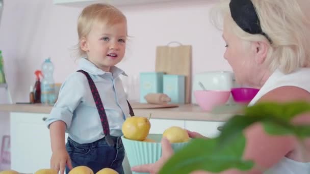 Porträt eines blonden kleinen Jungen, der einen Korb mit Birnen umdreht, eine fröhliche kaukasische Seniorin, die lacht. Charmantes Kind spielt mit Großmutter in der heimischen Küche. — Stockvideo