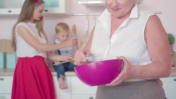 Eine bis zur Unkenntlichkeit glückliche Seniorin blickt zurück auf Tochter und Enkel, die spielen und in die Kamera lächeln. Fürsorgliche Großmutter bereitet zu Hause in der Küche Abendessen für Familie zu. — Stockvideo