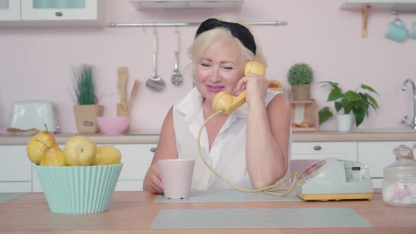 Szczęśliwa seniorka rozmawiająca przez telefon obrotowy siedząca przy stole w kuchni retro. Portret spokojnego, białego emeryta odpoczywającego w domu w latach 60-tych. Koncepcja czasu wolnego na emeryturze. — Wideo stockowe