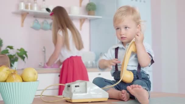 Uroczy, biały chłopczyk siedzący na stole i bawiący się obrotowym telefonem, jak rozmyta młoda kobieta zmywająca naczynia w tle. Portret szczęśliwej rodziny kaukaskiej z lat 60-tych w kuchni. — Wideo stockowe