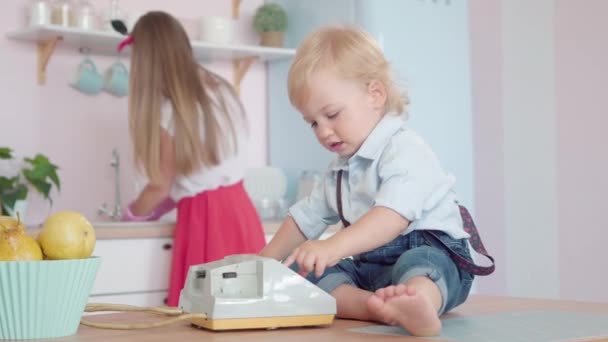 Rack focus change de jeune mère souriante laver la vaisselle à mignon petit garçon jouant avec téléphone rotatif. Heureux parent caucasien et enfant se reposant à la maison dans les années 1960. — Video