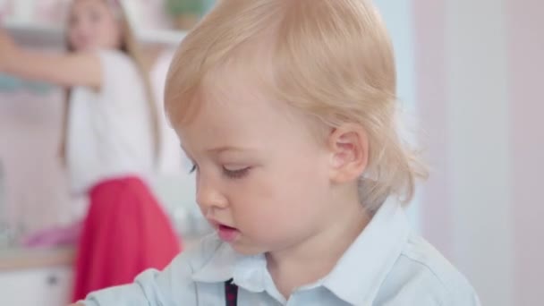 Gros plan de mignon garçon caucasien blond jouant avec téléphone rotatif comme femme floue laver la vaisselle à l'arrière-plan. Portrait de charmant bambin dans les années 1960. — Video