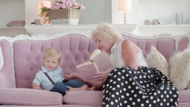 Positivo libro di lettura della nonna caucasica per il piccolo ragazzo biondo carino. Bambino annoiato che sale sul divano e se ne va. Bella donna anziana che parla col nipote e sorride. Buon fine settimana. — Video Stock