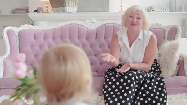Sød lille dreng giver buket lyserøde tulipaner til smilende senior kvinde sidder på sofaen. Blond kaukasisk lille barn lykønsker bedstemor med ferie. Barnebarn møde bedsteforældre. – Stock-video
