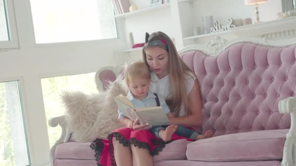 Joyeux livre de lecture de jeune femme pour mignon petit garçon assis sur le canapé à la maison. Portrait de charmante mère et fils caucasiens appréciant le temps ensemble à l'intérieur. Concept de famille. — Video