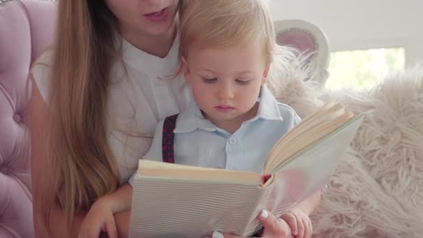 Gros plan d'un tout-petit concentré regardant dans le livre comme une mère méconnaissable lisant une histoire. Portrait de mignon petit garçon caucasien profitant week-ends avec jeune femme. Concept d'enfance heureuse. — Video