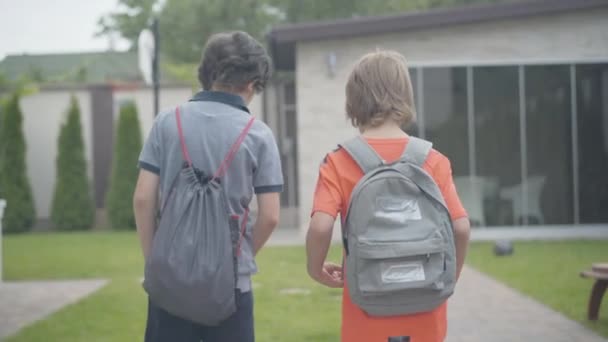 放課後、 2人の少年が帰宅する姿をバックに。白人の少年たちが家の庭を歩いて話している。教育と幼少期の概念. — ストック動画