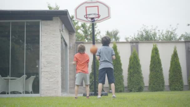 Två glada små pojkar som spelar basket hemma. Porträtt av glada avslappnade kaukasiska skolpojkar vilar på helgerna utomhus. Idrott och barndom. — Stockvideo
