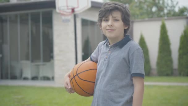 Portret de băiat brunetă încrezător pozând cu mingea de baschet în aer liber. Dragut caucazian scolar uitandu-se la camera si zambind. Un stil de viață sănătos și un concept sportiv . — Videoclip de stoc
