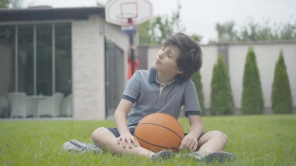 Menino caucasiano pensativo sentado na grama verde com bola como irmão borrado escalando no aro de basquete no fundo. Retrato de criança bonito sonhando com carreira esportiva ao ar livre. — Vídeo de Stock