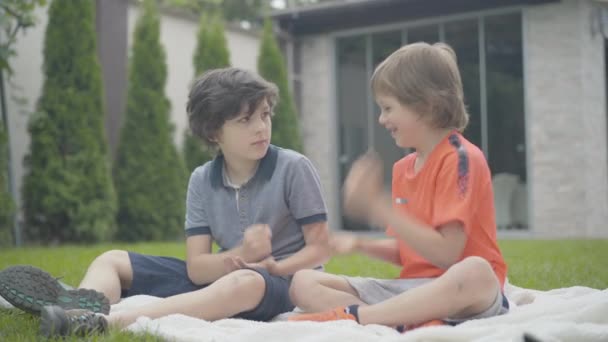 Brede opname van zorgeloze jongens die buiten steen-papier-schaar spelen. Portret van twee vrolijke blanke schooljongens die op een deken zitten en praten. Vrienden hebben plezier in het weekend. — Stockvideo