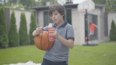 Küçük beyaz bir çocuk topuyla ferahlatıcı meyve suyu içiyor arka plandaki basketbol potasına tırmanan bulanık bir öğrenci gibi. Rahatlamış kaygısız çocuklar hafta sonları dışarıda eğleniyor..