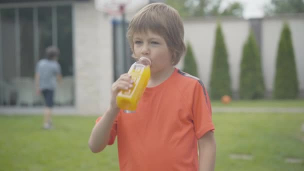 Portret szczęśliwego białego chłopca pijącego żółty orzeźwiający sok i uśmiechającego się do kamery. Wesoły dzieciak ze zdrowym drinkiem na świeżym powietrzu. Niewyraźny brat lub przyjaciel grający w koszykówkę w tle. — Wideo stockowe