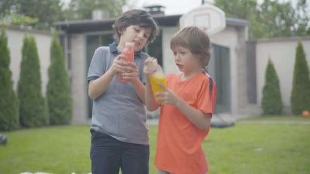 Engraçado meninos tremendo suco colorido em garrafas e falando. Retrato de amigos caucasianos despreocupados se divertindo nos fins de semana ao ar livre. Conceito de infância e lazer. — Vídeo de Stock